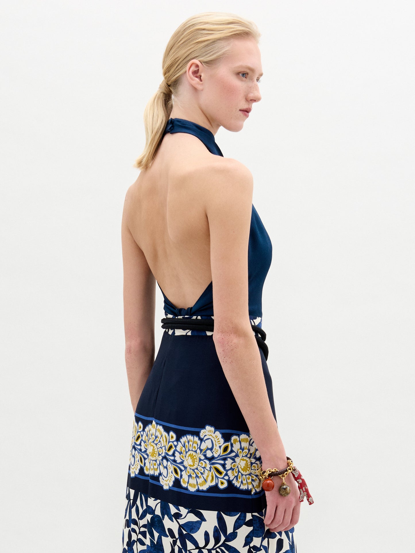 Someone with blond hair in a low ponytail wears the SVT DESIGNS Colima Top, crafted from exquisite Italian fabric. The navy halter top features an exposed back and shoulders, with a floral design at the bottom and a patterned belt. A colorful beaded bracelet complements the look against a plain backdrop.