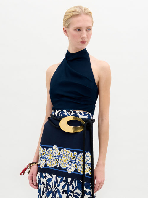 A person with blonde hair stands against a plain background, wearing the sleeveless Colima Top by SVT DESIGNS, crafted from luxurious Italian fabric. They also sport a patterned skirt with floral designs and a prominent gold belt, gazing to the side.