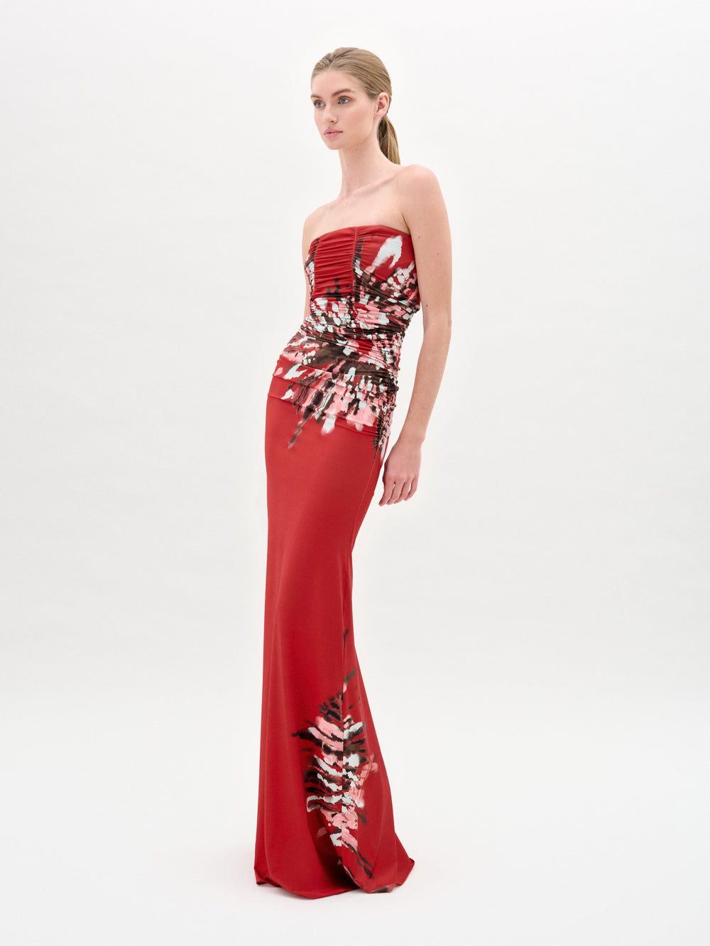 A woman wears the SVT DESIGNS Collins Dress, a strapless maxi crafted from luxurious Italian fabric with a fitted bodice. The floor-length red dress features an abstract pattern of pink, white, and black against a plain white background.