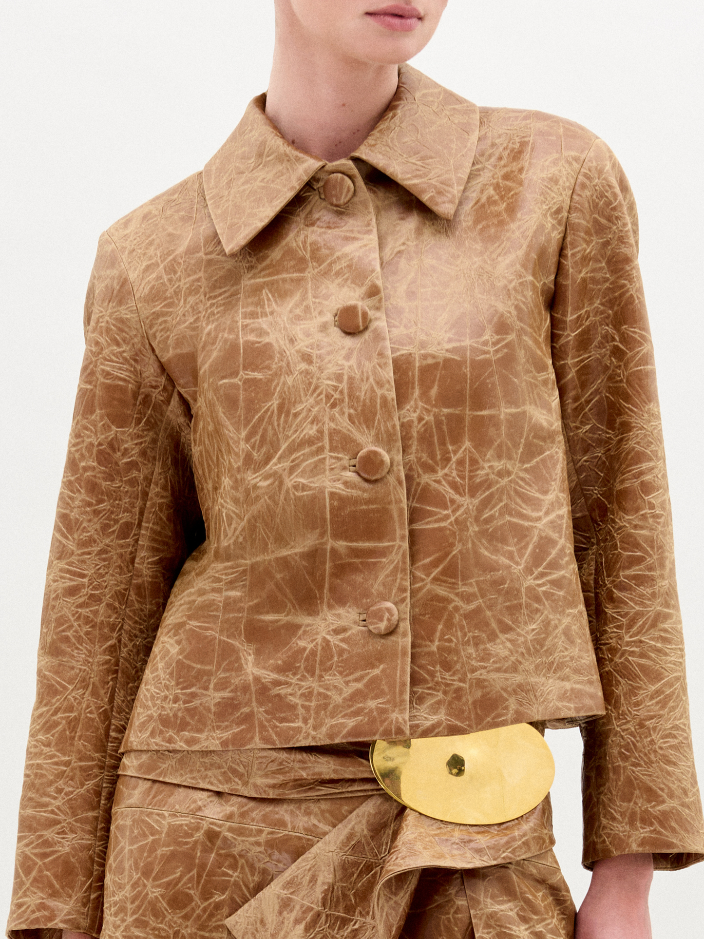 A woman wearing the Columba Jacket—a brown, textured, structured jacket with a classic collar—and a matching skirt with a gold belt buckle stands against a plain white background.