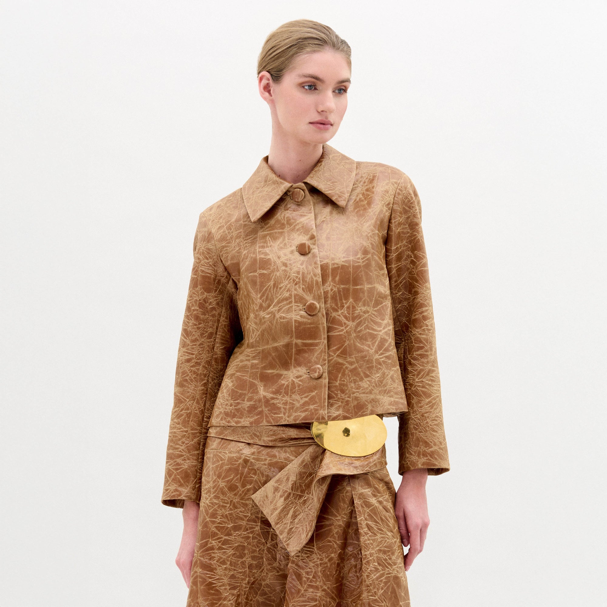 A woman wearing the Columba Jacket—a brown, textured, structured jacket with a classic collar—and a matching skirt with a gold belt buckle stands against a plain white background.