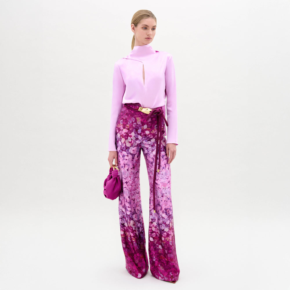 A person stands against a white background wearing SVT DESIGNS' Como Pant—high-waisted, wide-leg Italian fabric pants in purple and pink print—with a light pink slit-sleeve top and holding a matching purple handbag.