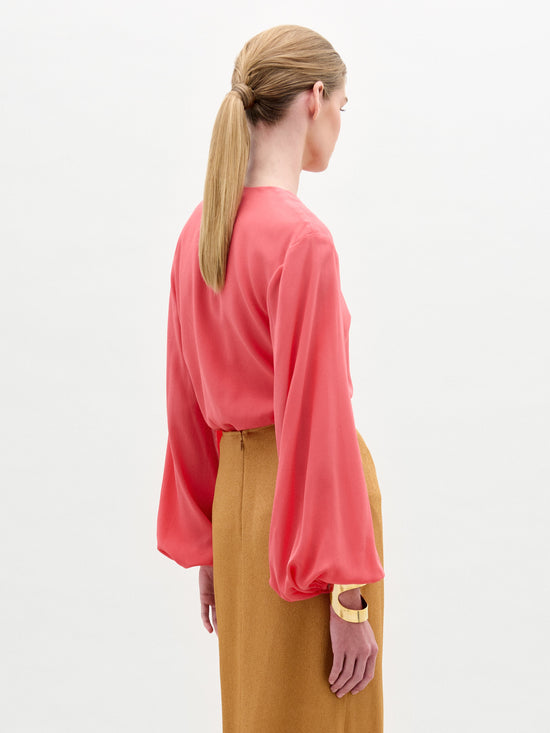 A person with long hair in a low ponytail wears the Covilha Blouse by SVT DESIGNS, a coral, long-sleeved, loose-fitting top crafted from Italian fabric. They pair it with a high-waisted mustard skirt and glance over their left shoulder against a plain, light-colored background.