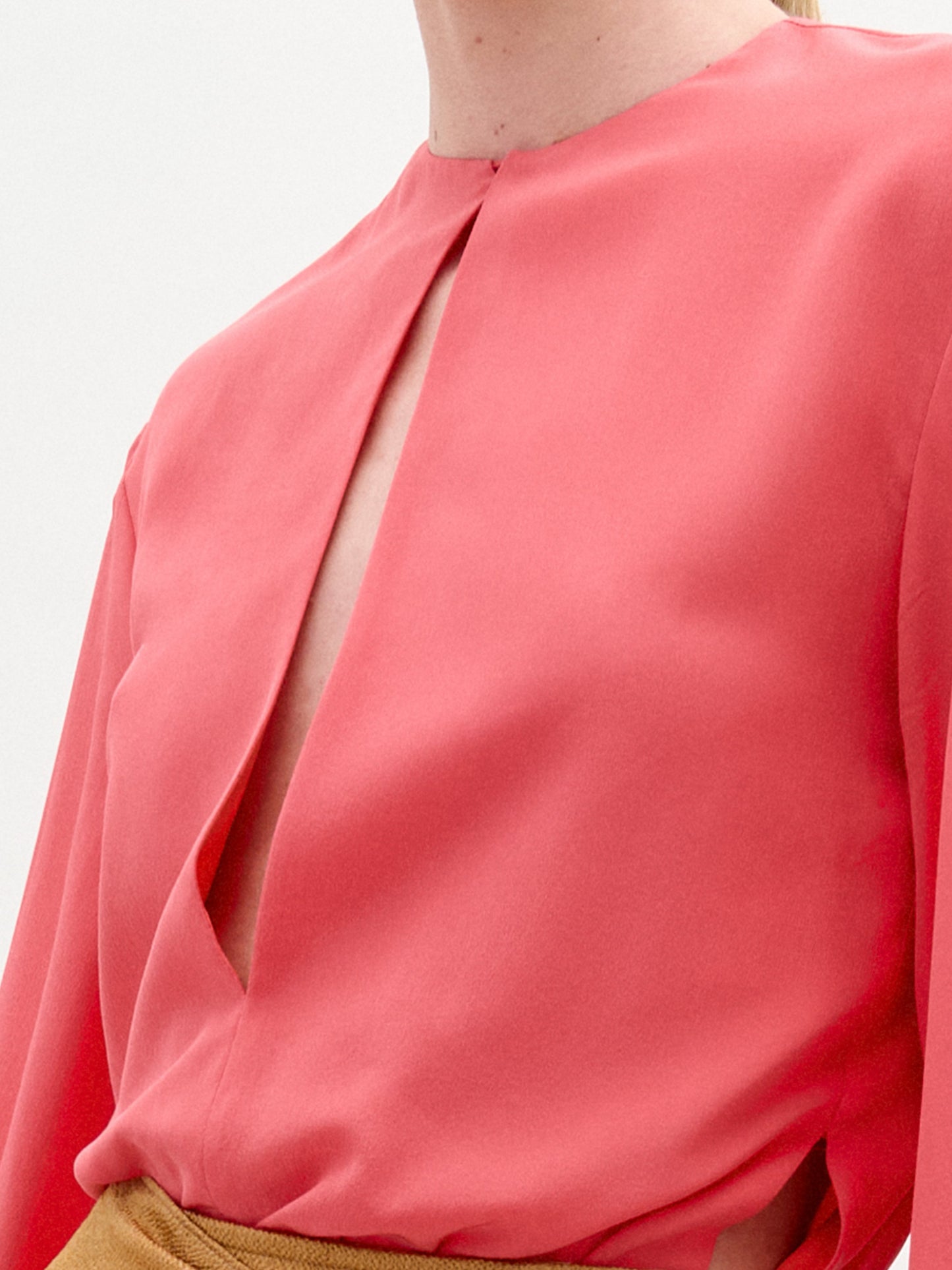 A close-up showcases a person wearing the Covilha Blouse by SVT DESIGNS, a pink 100% silk piece with a front slit design. Crafted from Italian fabric, this long-sleeve blouse is paired with a brown skirt and set against a plain white background, focusing on the upper portion.