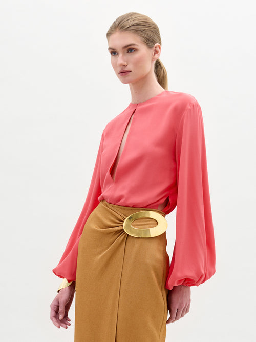 A woman stands against a plain background, wearing the Covilha Blouse by SVT DESIGNS, a coral long-sleeve blouse made from luxurious Italian fabric, paired with a tan skirt featuring a large gold buckle. Her hair is styled in a low ponytail.