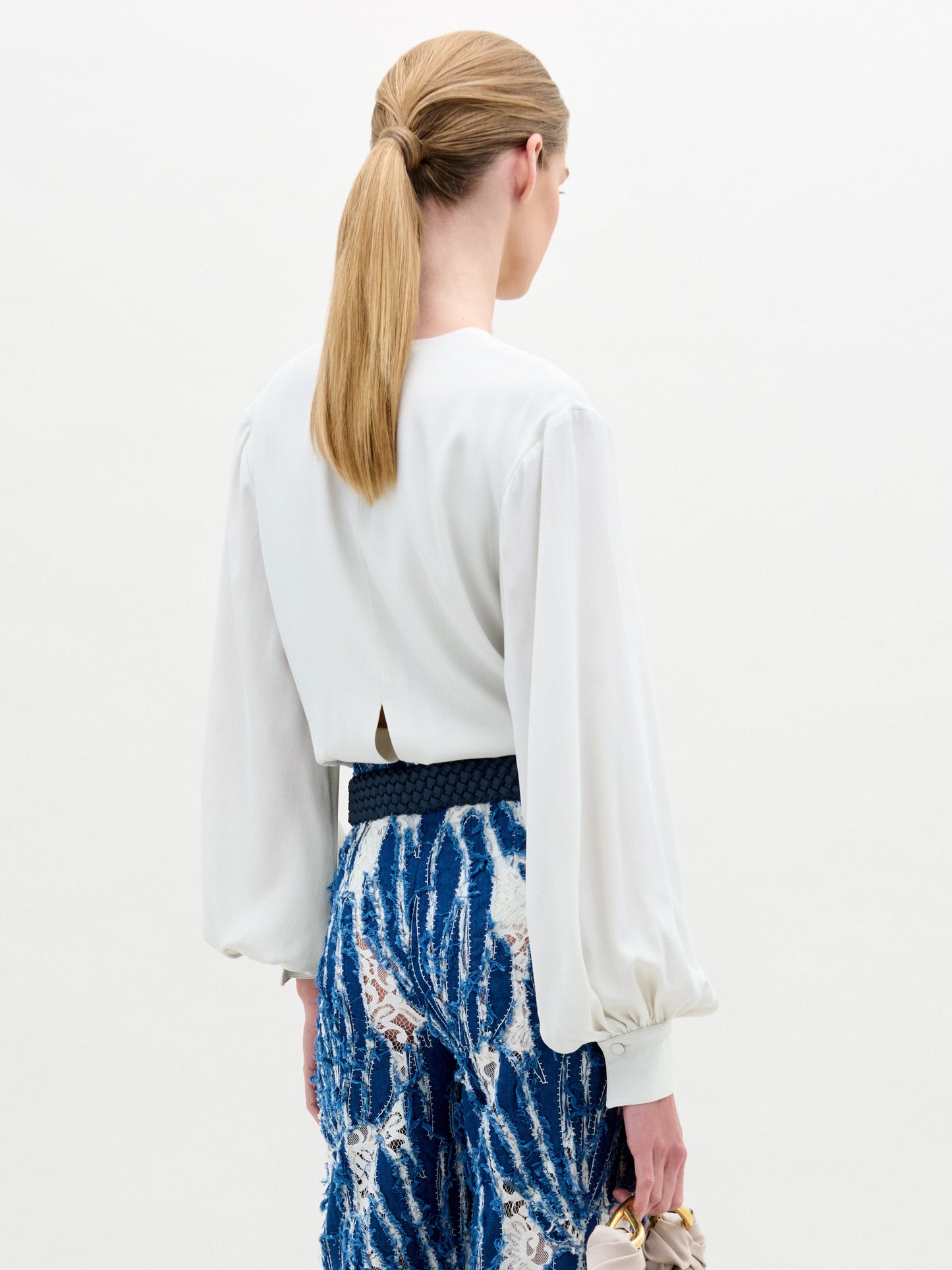 A person with blond ponytail wears SVT DESIGNS' Covilha Blouse, a white silk top featuring a keyhole neckline, paired with a blue lace skirt detailed in white and black. Holding a beige handbag, they stand against a plain background facing away.