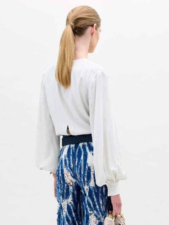 A person with blond ponytail wears SVT DESIGNS' Covilha Blouse, a white silk top featuring a keyhole neckline, paired with a blue lace skirt detailed in white and black. Holding a beige handbag, they stand against a plain background facing away.