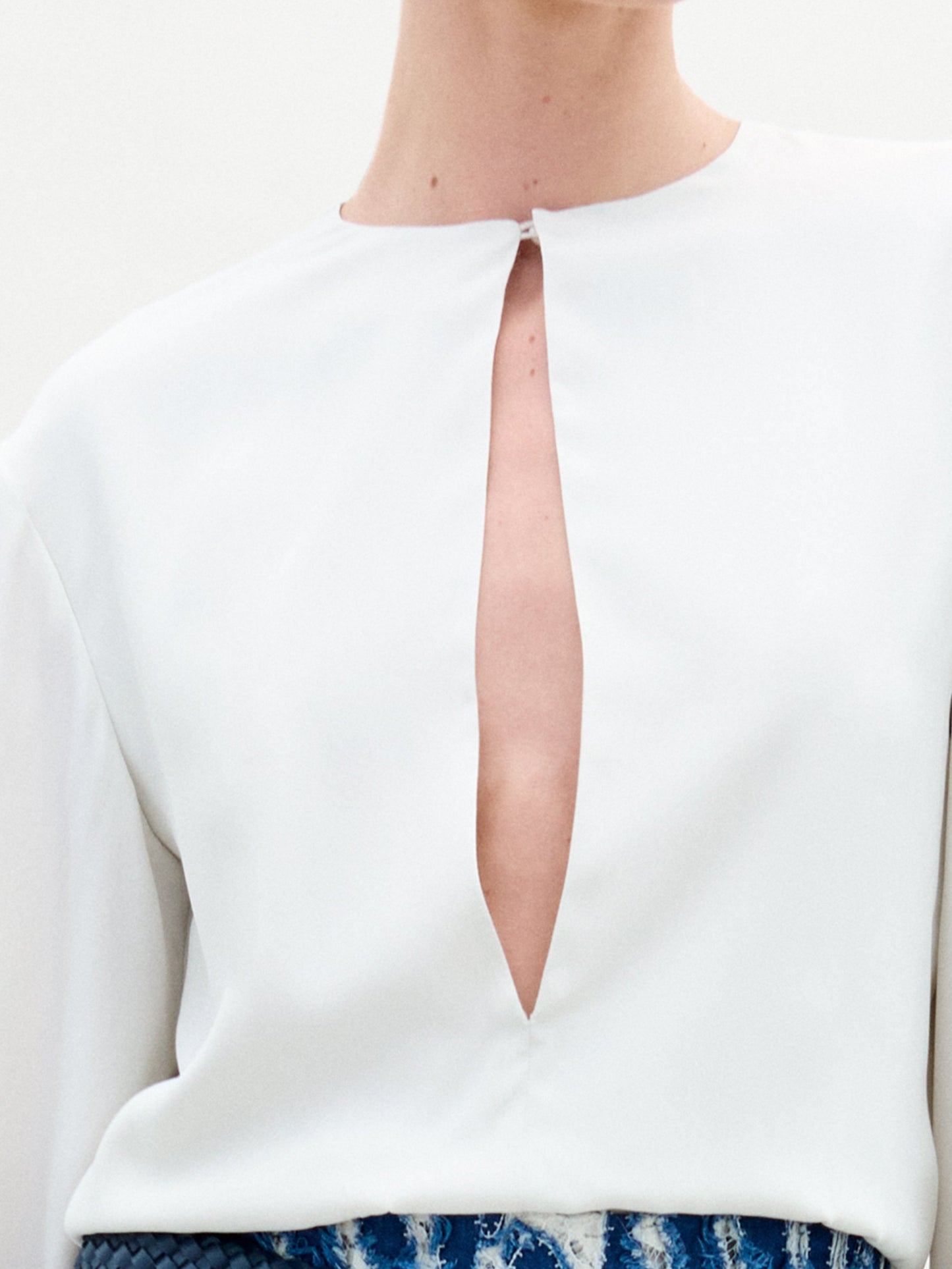 A close-up shows a person wearing the Covilha Blouse by SVT DESIGNS, featuring a V-neckline and long sleeves crafted from fine Italian silk. The smooth white garment overlays a textured blue and white piece beneath, focusing on the upper chest area while keeping the face unseen.