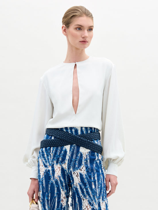 A person is wearing the Covilha Blouse by SVT DESIGNS, a white silk blouse with a keyhole neckline, and blue patterned pants with a textured blue belt. The background is plain white.