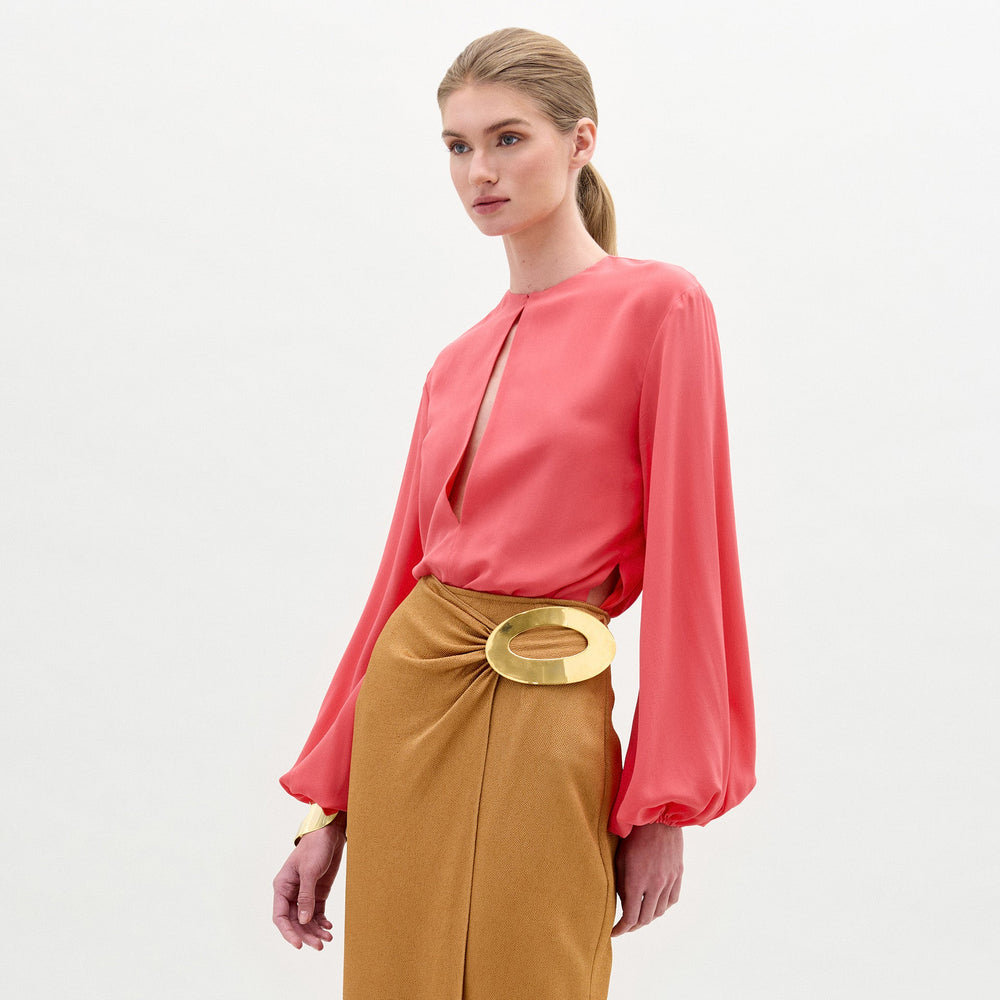 A woman stands against a plain light background wearing the SVT DESIGNS Covilha Blouse in coral with a keyhole neckline and an ochre skirt with a large gold circular belt. Her hair is tied back in a low ponytail.