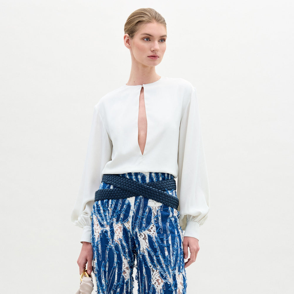 Wearing the SVT DESIGNS Covilha Blouse—a white silk top with a keyhole neckline and long balloon sleeves—paired with high-waisted blue patterned pants, and holding a small bag against a plain background.