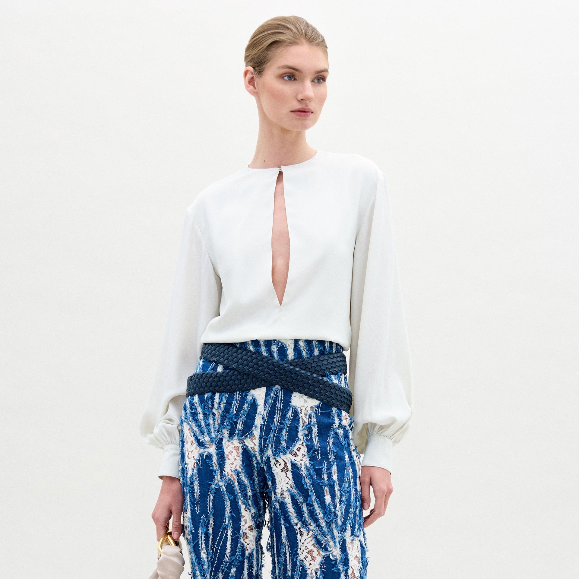 Wearing the SVT DESIGNS Covilha Blouse—a white silk top with a keyhole neckline and long balloon sleeves—paired with high-waisted blue patterned pants, and holding a small bag against a plain background.