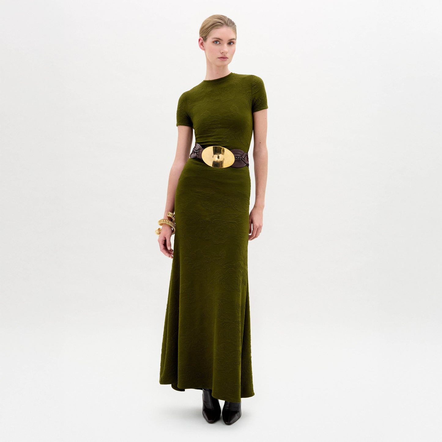 A woman wearing the Cristal Dress, an olive green slim fit maxi dress paired with a wide black belt with a gold buckle, black boots, and gold bracelets stands against a plain white background.