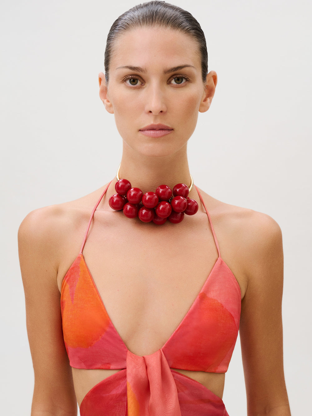 A woman wears the Candida Dress, a slim-fit, halter-neck style in shades of orange and pink, accessorized with a large, clustered red beaded necklace.