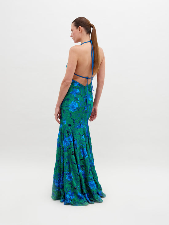 A person stands wearing a vibrant green and blue floral-patterned dress with an open back and a halter neck, posing against a plain white background. The Cara Dress Green Navy Embroidery is available for pre-order now, with a ship by date of November 15th, 2024.