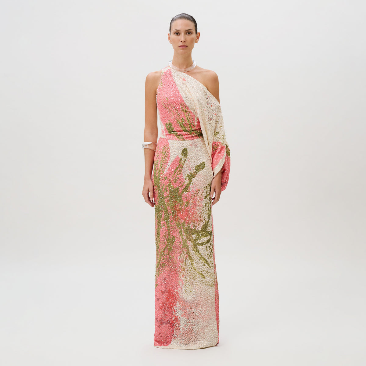 A model wears the Carmel Dress, a floor-length, one-shoulder gown made in Colombia, featuring an abstract pink and olive green pattern woven with sequins and a draped sleeve.