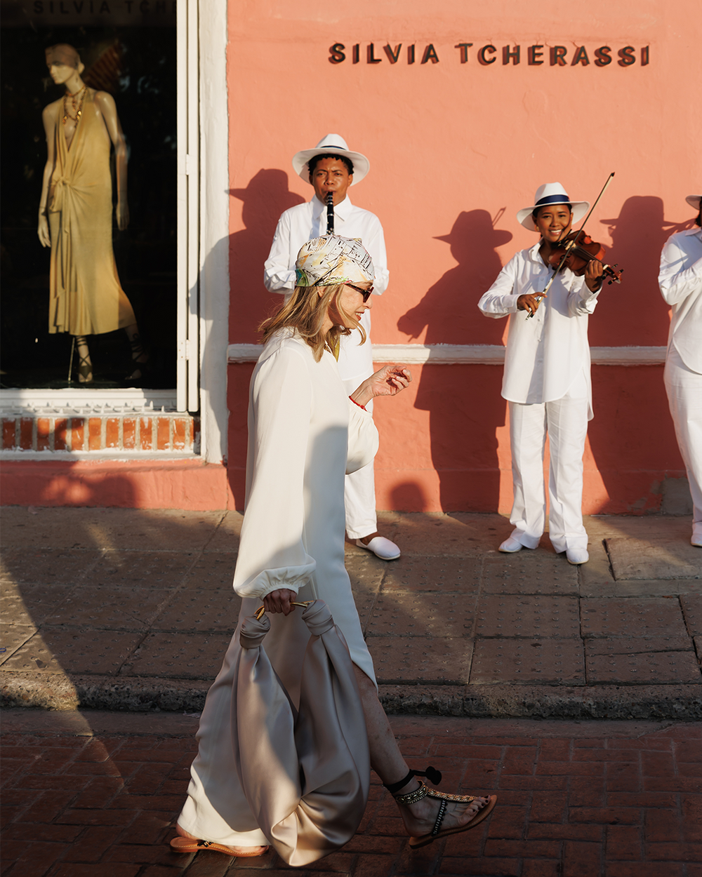 Silvia Tcherassi in a white dress walking past a store with mannequins and a pink wall.
