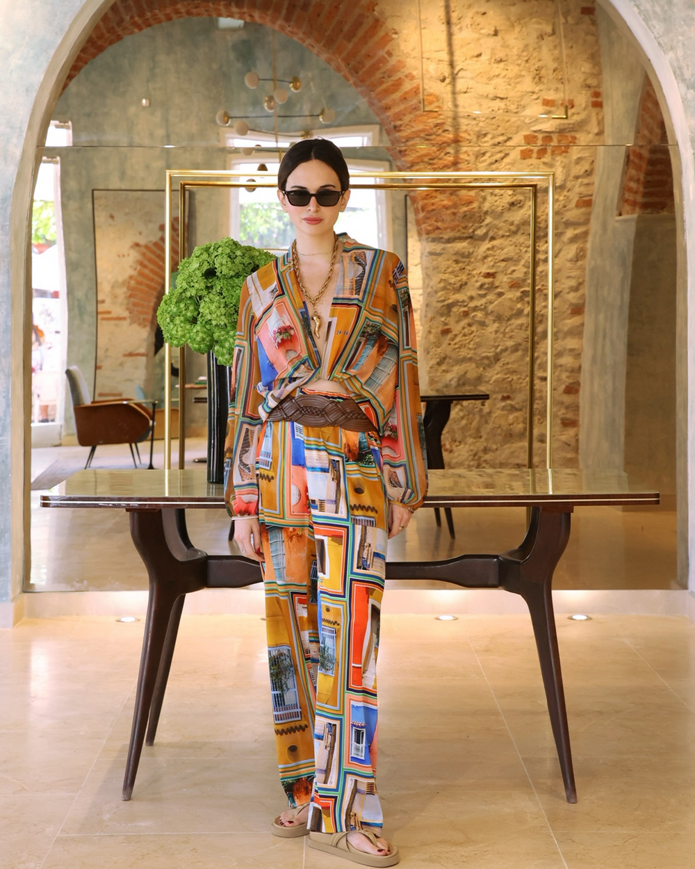 Woman in a colorful outfit standing in an elegant interior setting with mirrors and a table.