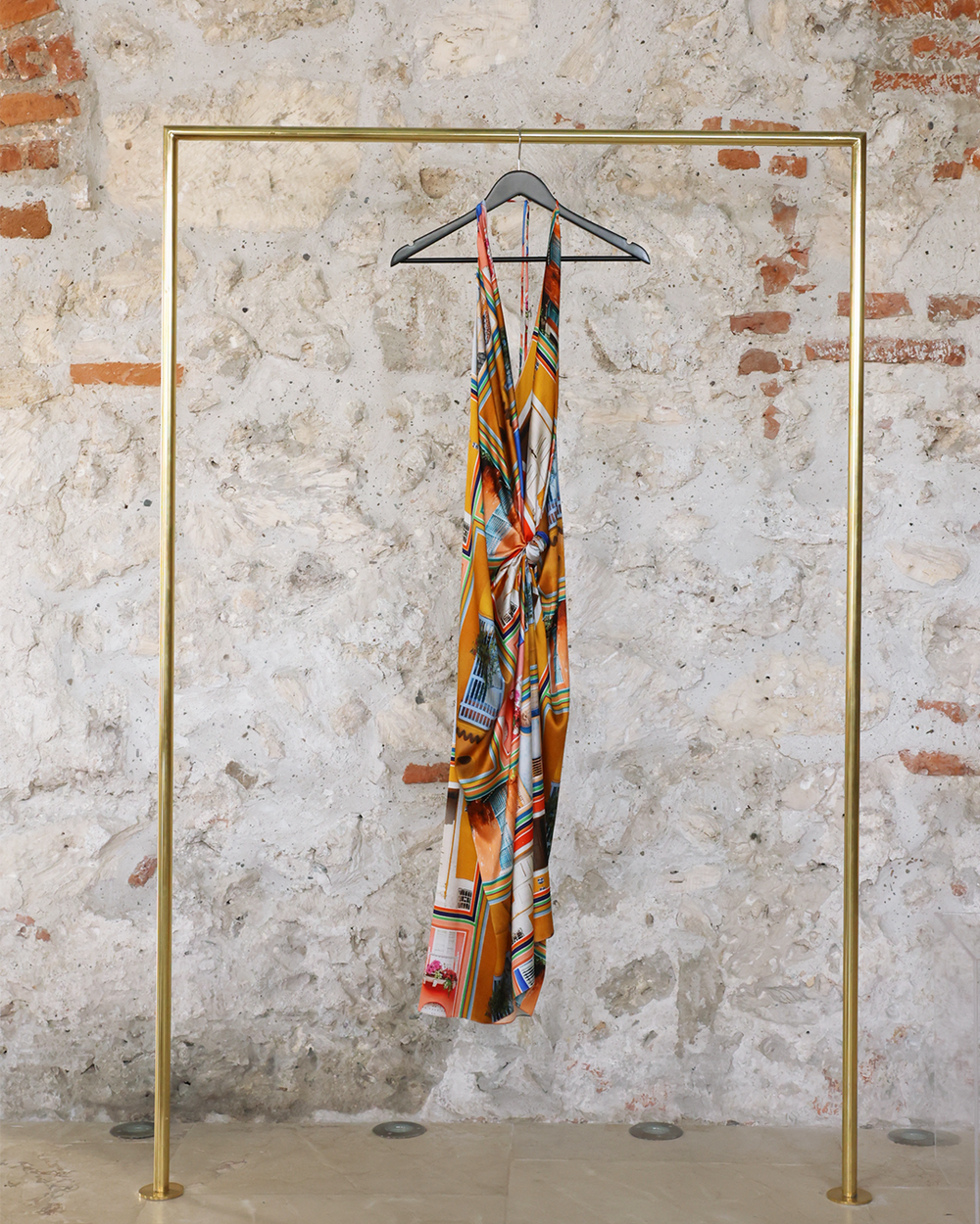 Abstract printed halter dress hanging on a minimalist gold rack against a textured stone wall background.