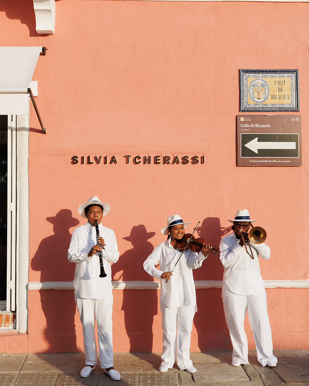 Three musicians in white outfits playing instruments against a pink wall with 'Silvia Tcherassi' branding.