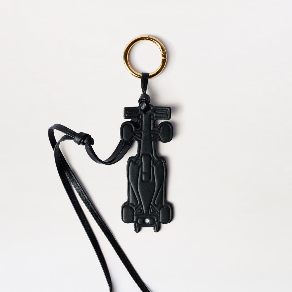A black leather keychain shaped like a Formula 1 race car, featuring a gold-tone split keyring and a black leather strap, shown on a clean white background.