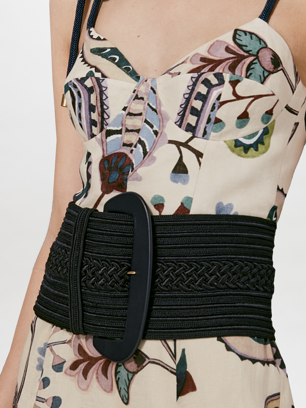A close-up of a person wearing a patterned sleeveless dress cinched at the waist with the Silvia Tcherassi US Chenoa Belt, featuring a wide black fabric and a large oval buckle. The soft viscose dress displays muted floral and geometric designs.