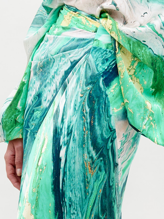 A person wearing the Claudia Skirt Green Marble dress, featuring a long-sleeved design with a shoulder cutout, poses against a plain white background. Pre-order now to ship by February 15th, 2025.