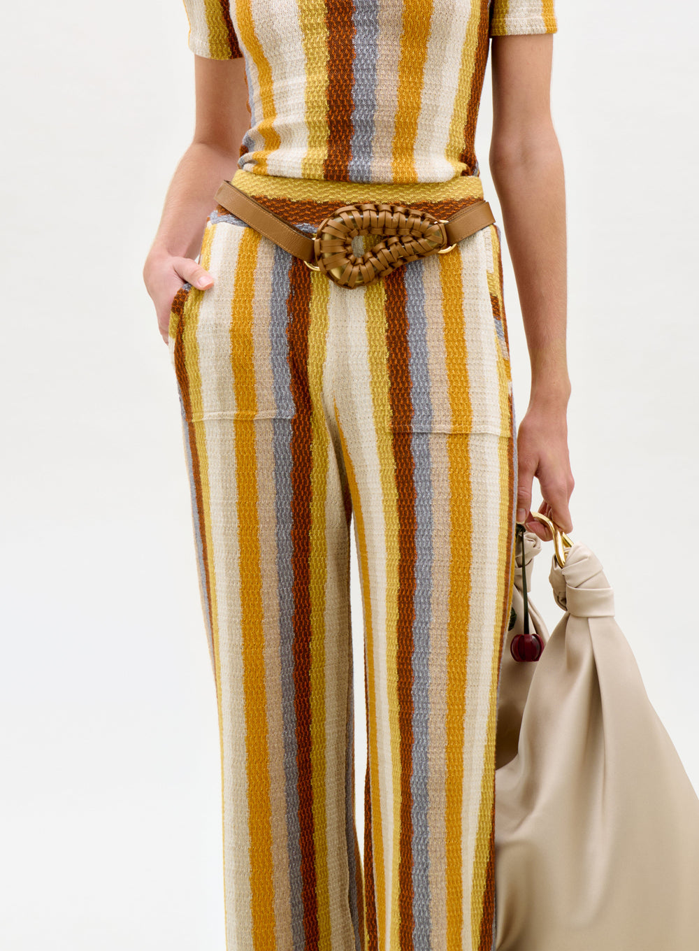 Person wearing a striped outfit with a belt and holding a beige bag on a white background