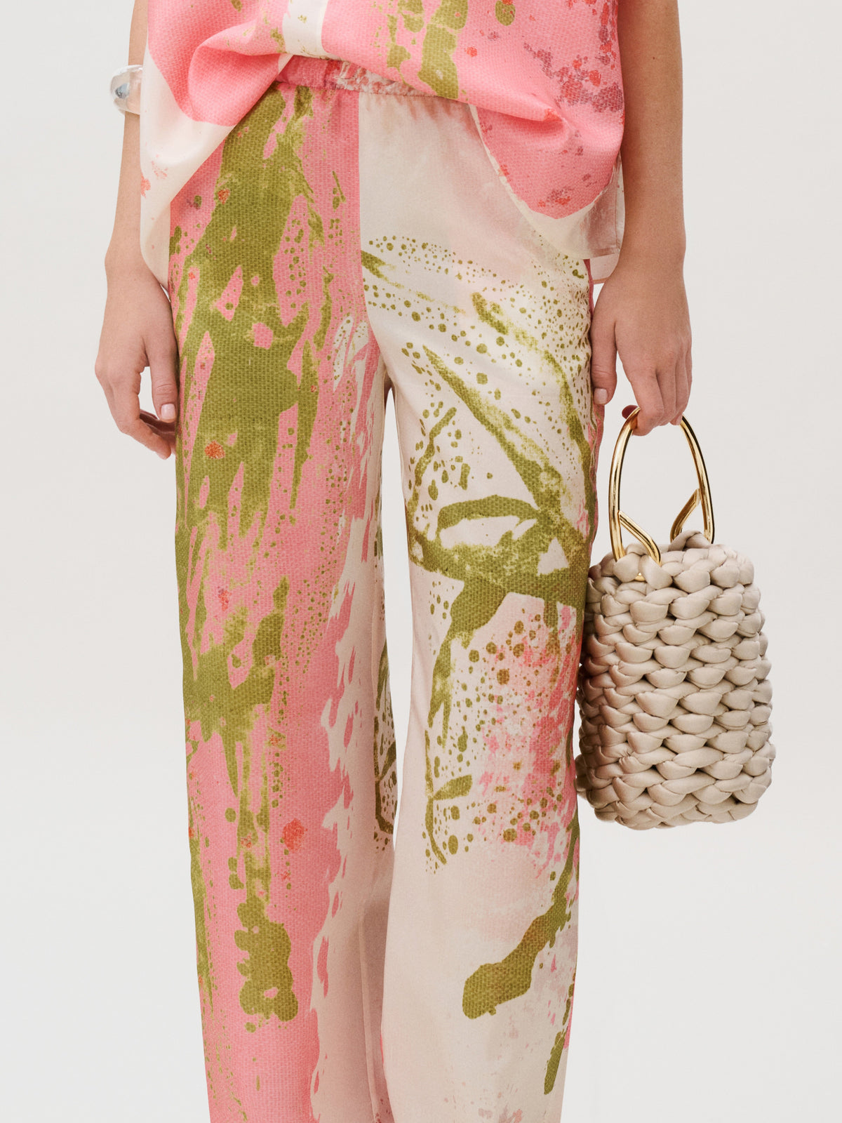 Curacao Pant in Italian silk habutai with a pink and green abstract paint splatter print, worn with a woven cream handbag with gold circular handles.