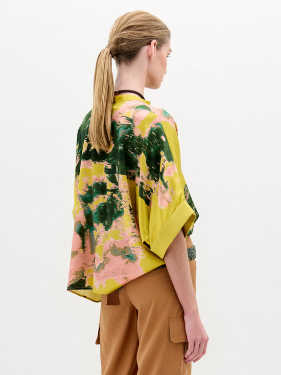 A person with long hair in a ponytail, seen from the side, wears an SVT DESIGNS Daia Blouse—an oversized fit, tie-dye kimono-style top crafted from Italian fabric in green, yellow, and pink. They're also wearing tan pants and a black necklace against a neutral background.