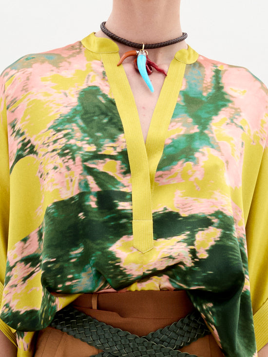 A person wears the Daia Blouse by SVT DESIGNS, an oversized fit with a yellow and green abstract pattern and V-neck, crafted from luxurious Italian fabric. They accessorize with a brown woven belt and a brown choker necklace featuring a blue and red pendant.