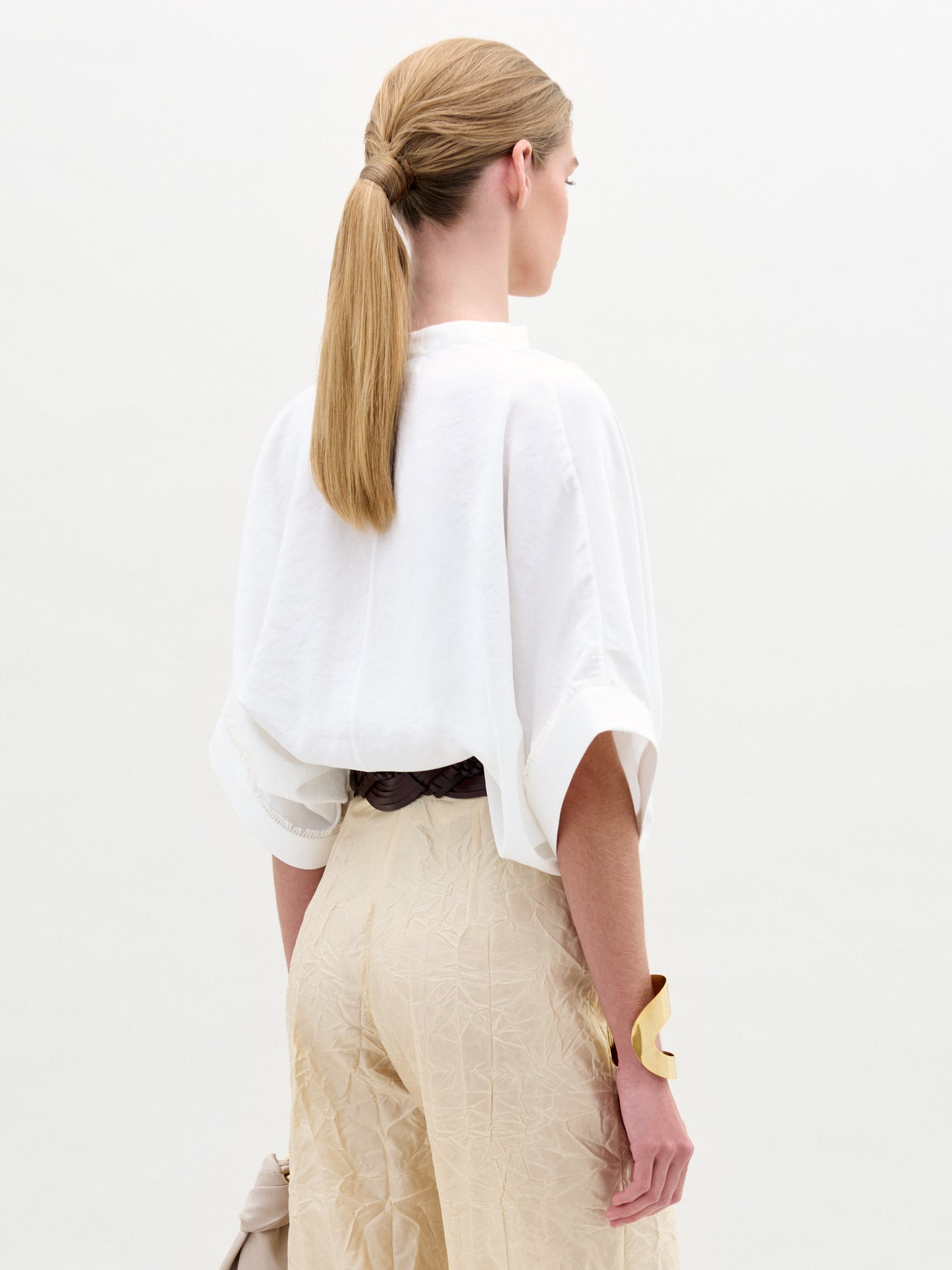 A person with long, straight hair tied back is facing away. They're wearing an oversized Daia Blouse by SVT DESIGNS with a V-cut neckline and rolled sleeves, paired with beige high-waisted pants. An Italian fabric adds sophistication, and a large gold bracelet stands out against the plain white background.