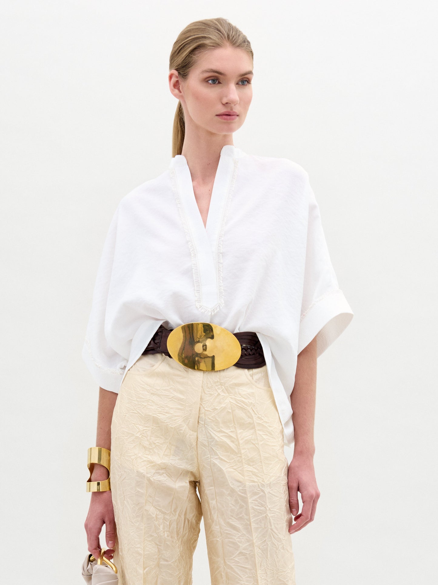 A woman models the oversized Daia Blouse by SVT DESIGNS, featuring a loose white top with a V-cut neckline and beige pants made from fine Italian fabric. She accessorizes with gold bracelets and a large gold belt buckle against a plain white background.