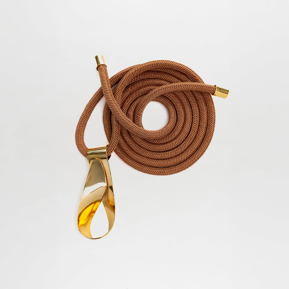 The Daiana Belt is a coiled brown cord belt featuring gold metal end caps and a polished gold-plated brass buckle, made from fine Italian material and shown on a white background.