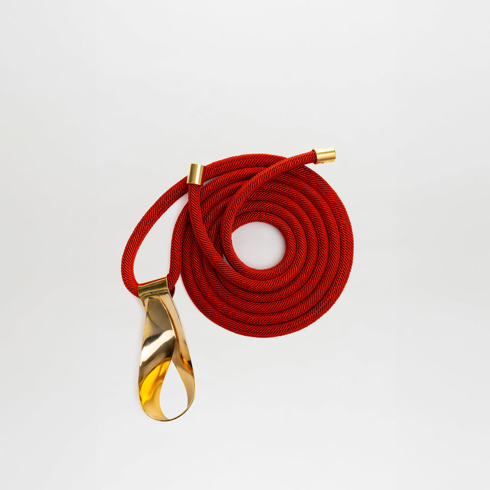 The Daiana Belt by SVT DESIGNS features a coiled red cord with gold metal caps and a large gold-plated brass buckle on one end, shown against a plain white background.