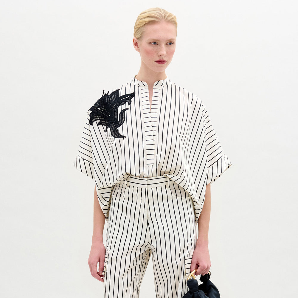 A person wears the Daia Blouse by SVT DESIGNS— a black-and-white vertical striped, short-sleeved top with a high neckline and black floral embellishment on one shoulder, made from Italian fabric. Plain white background.