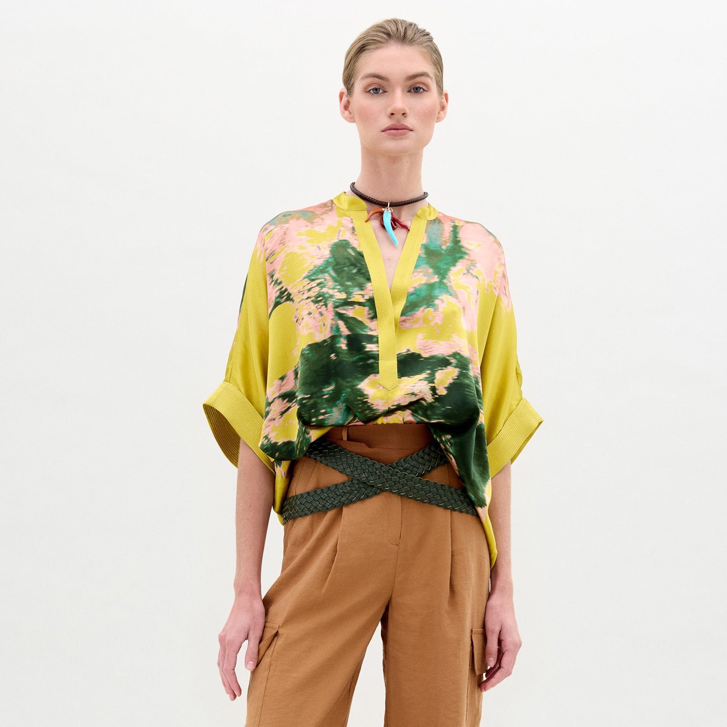 A person in the Daia Blouse by SVT DESIGNS—an oversized yellow, green, and pink floral piece with rolled sleeves made from Italian fabric—pairs it with tan belted pants and a choker necklace, standing against a plain white background.