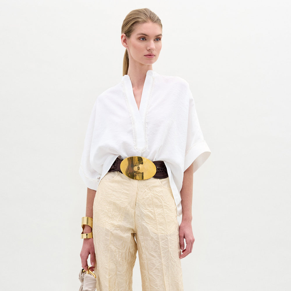 A person models the SVT DESIGNS Daia Blouse—a loose white top with wide sleeves and a V-neck—paired with cream textured pants, a wide belt with a large gold buckle, gold bracelet, and a small beige handbag against a plain white background.
