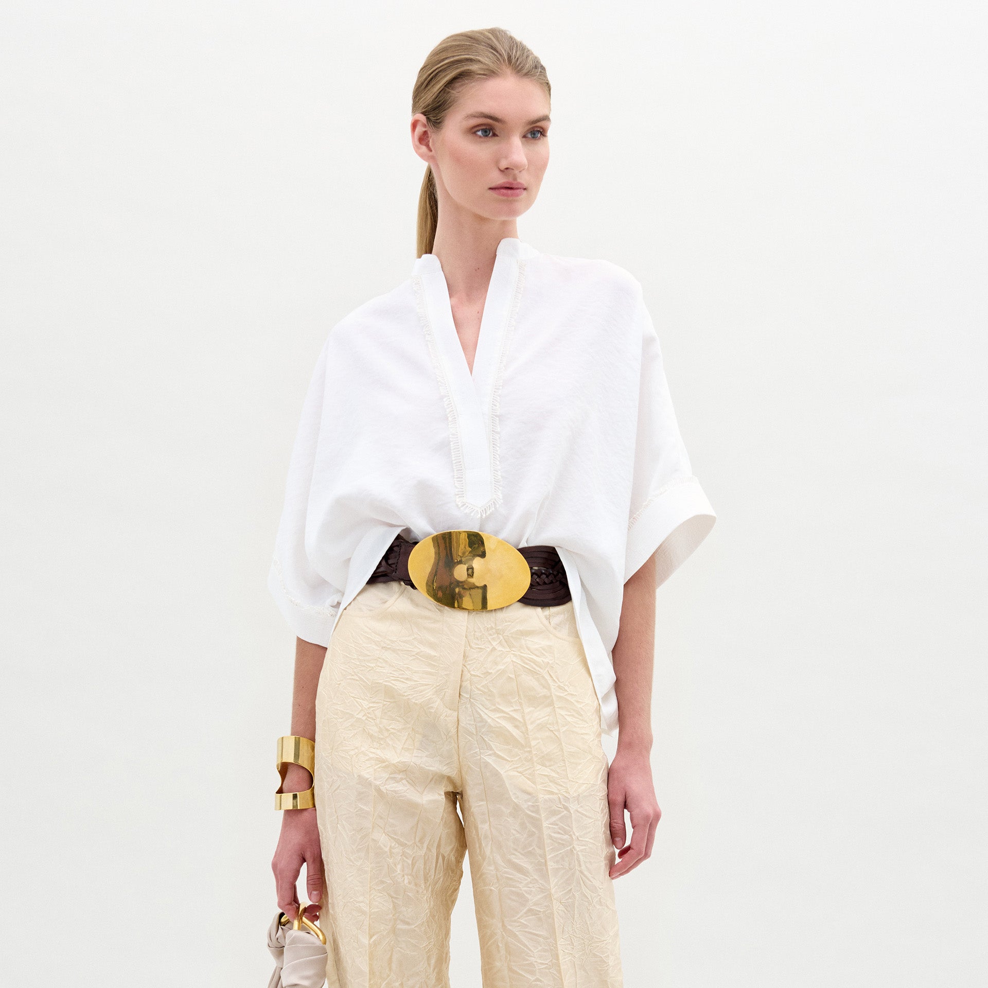 A person models the SVT DESIGNS Daia Blouse—a loose white top with wide sleeves and a V-neck—paired with cream textured pants, a wide belt with a large gold buckle, gold bracelet, and a small beige handbag against a plain white background.