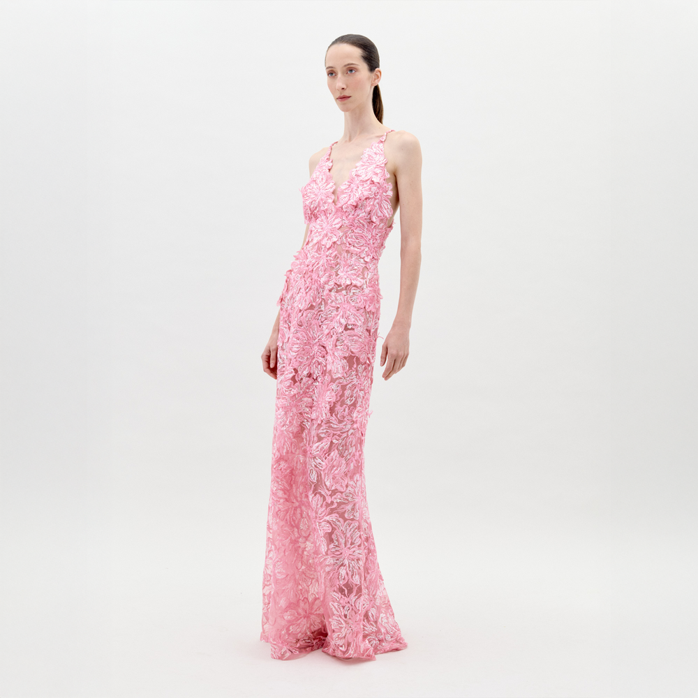 A woman wears the Daira Dress, a long, sleeveless pink tulle gown featuring intricate floral lace and delicate sequin embellishments, set against a plain light background.