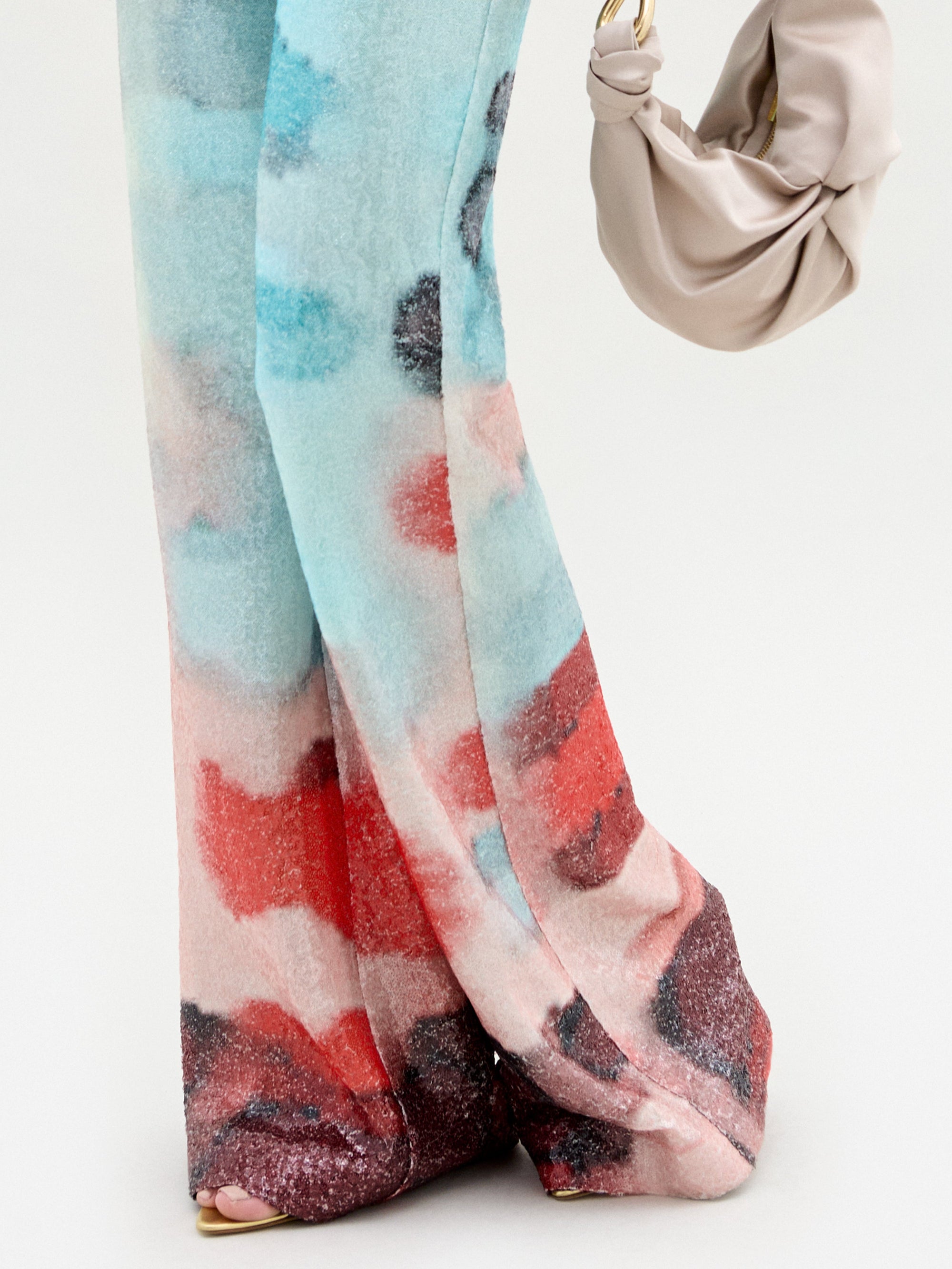 A person stands on a light surface, showing only the lower half, wearing SVT DESIGNS' Dalal Pant—high-waisted, wide-leg with a blue, red, pink, and black watercolor pattern—and holding a beige ruched handbag with a gold ring handle.