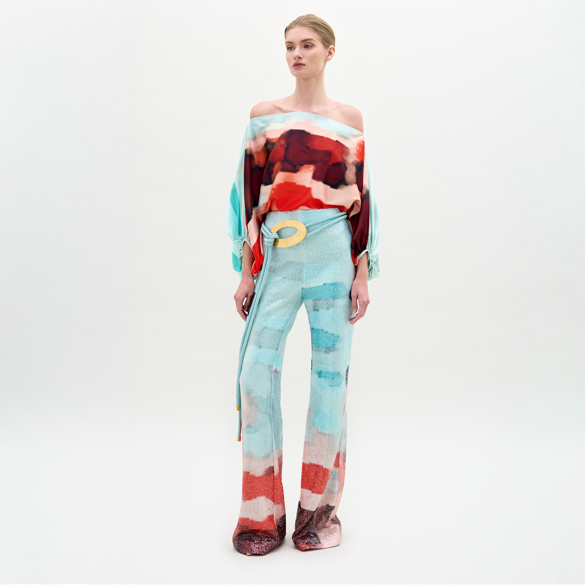 A person stands against a plain white background wearing SVT DESIGNS' Dalal Pant—high-waisted, wide-legged, with red and aqua abstract patterns—paired with a matching off-the-shoulder top and a light yellow belt.