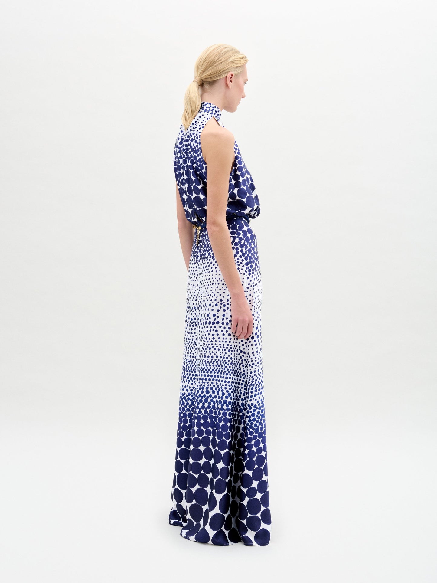 A blonde-haired individual stands sideways, wearing the Damara Dress from SVT DESIGNS. The sleeveless maxi dress, crafted from Italian fabric, showcases a blue and white dot pattern against a plain white background.