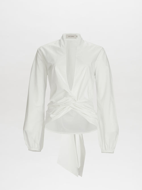 The Dania Blouse by SVT DESIGNS is a white long-sleeve crafted from 100% Italian cotton. It features a deep V-neckline, twisted knot detail at the front for a draped effect, and a back-tied sash. The fabric is smooth and lightweight.