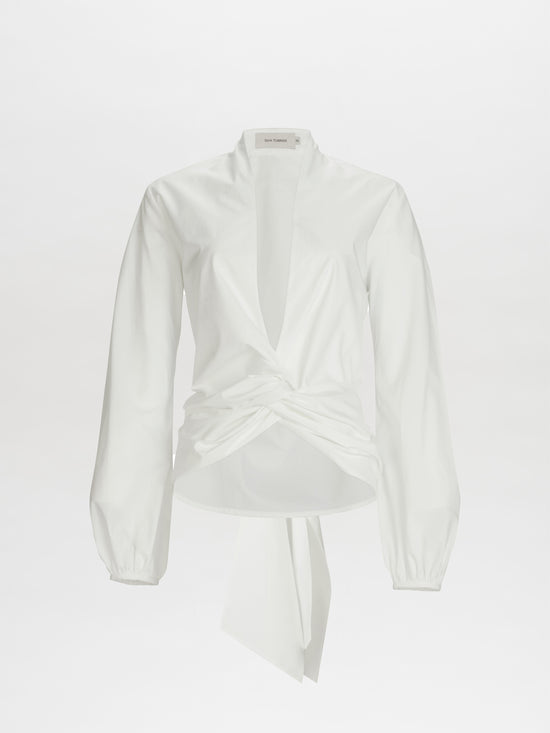 The Dania Blouse by SVT DESIGNS is a white long-sleeve crafted from 100% Italian cotton. It features a deep V-neckline, twisted knot detail at the front for a draped effect, and a back-tied sash. The fabric is smooth and lightweight.