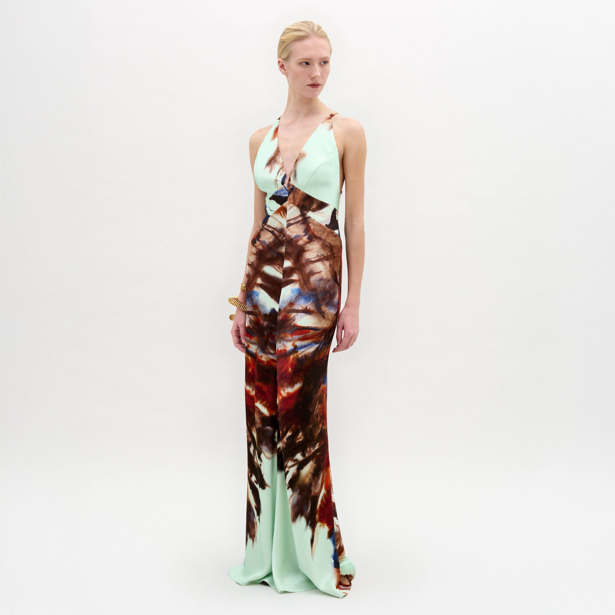 A woman poses in the SVT DESIGNS Daniella Dress, a sleeveless Italian-fabric maxi with a deep V-neckline. The mint green floor-length dress showcases a bold brown and red abstract pattern on the lower half.