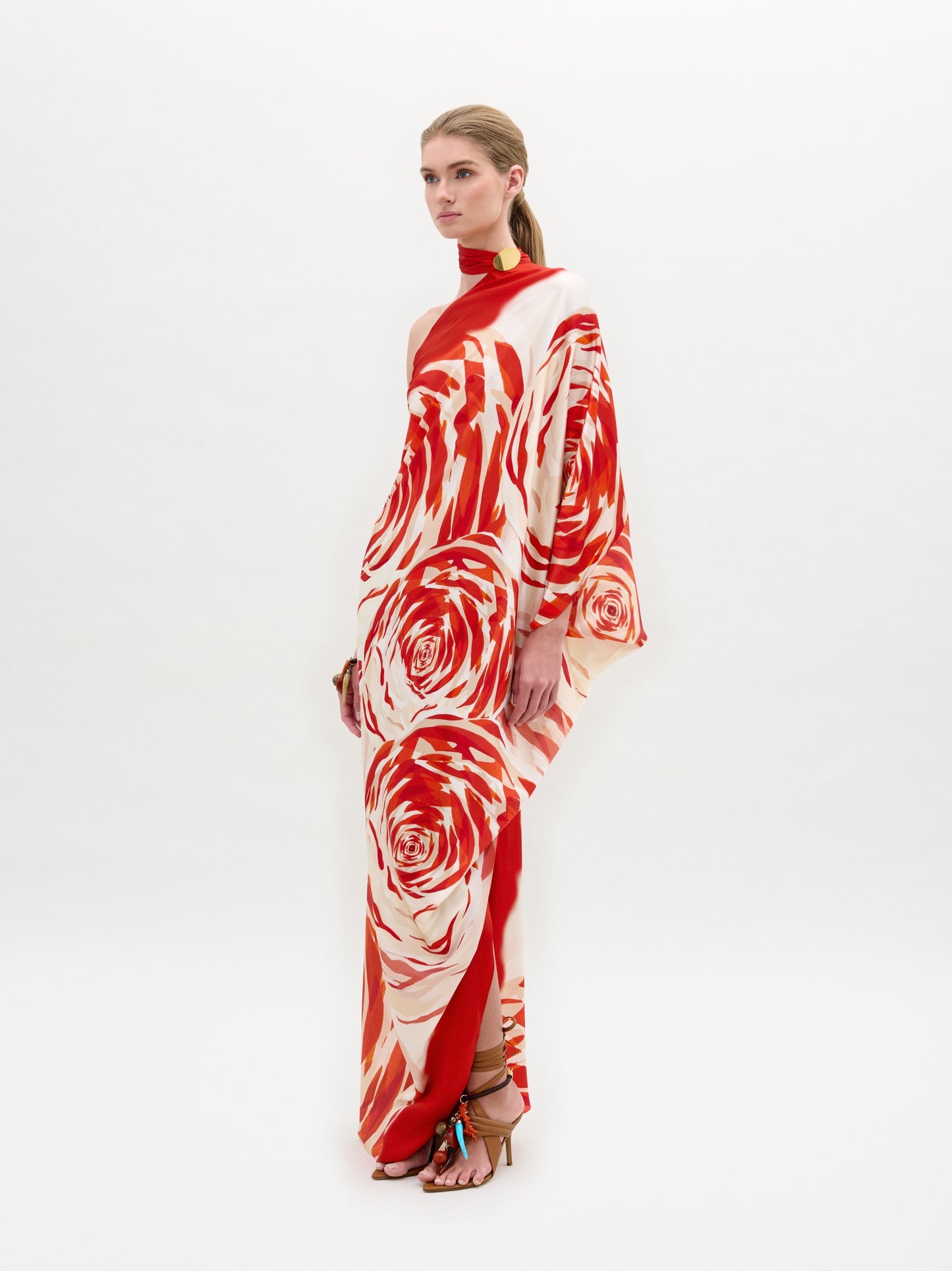 A person wearing the Dani Tunic by SVT DESIGNS stands confidently. It's a long, flowing silk maxi dress with red and white floral patterns, featuring one sleeve, a high neck, and a subtle side slit. Their long hair is tied back as they wear high-heeled sandals against a plain white background.