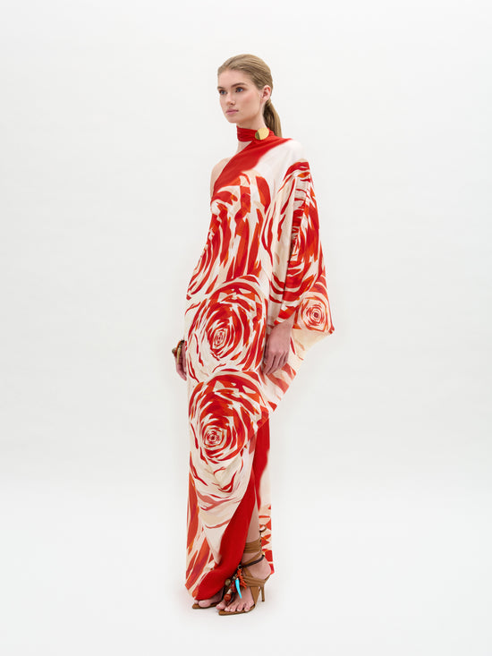 A person wearing the Dani Tunic by SVT DESIGNS stands confidently. It's a long, flowing silk maxi dress with red and white floral patterns, featuring one sleeve, a high neck, and a subtle side slit. Their long hair is tied back as they wear high-heeled sandals against a plain white background.