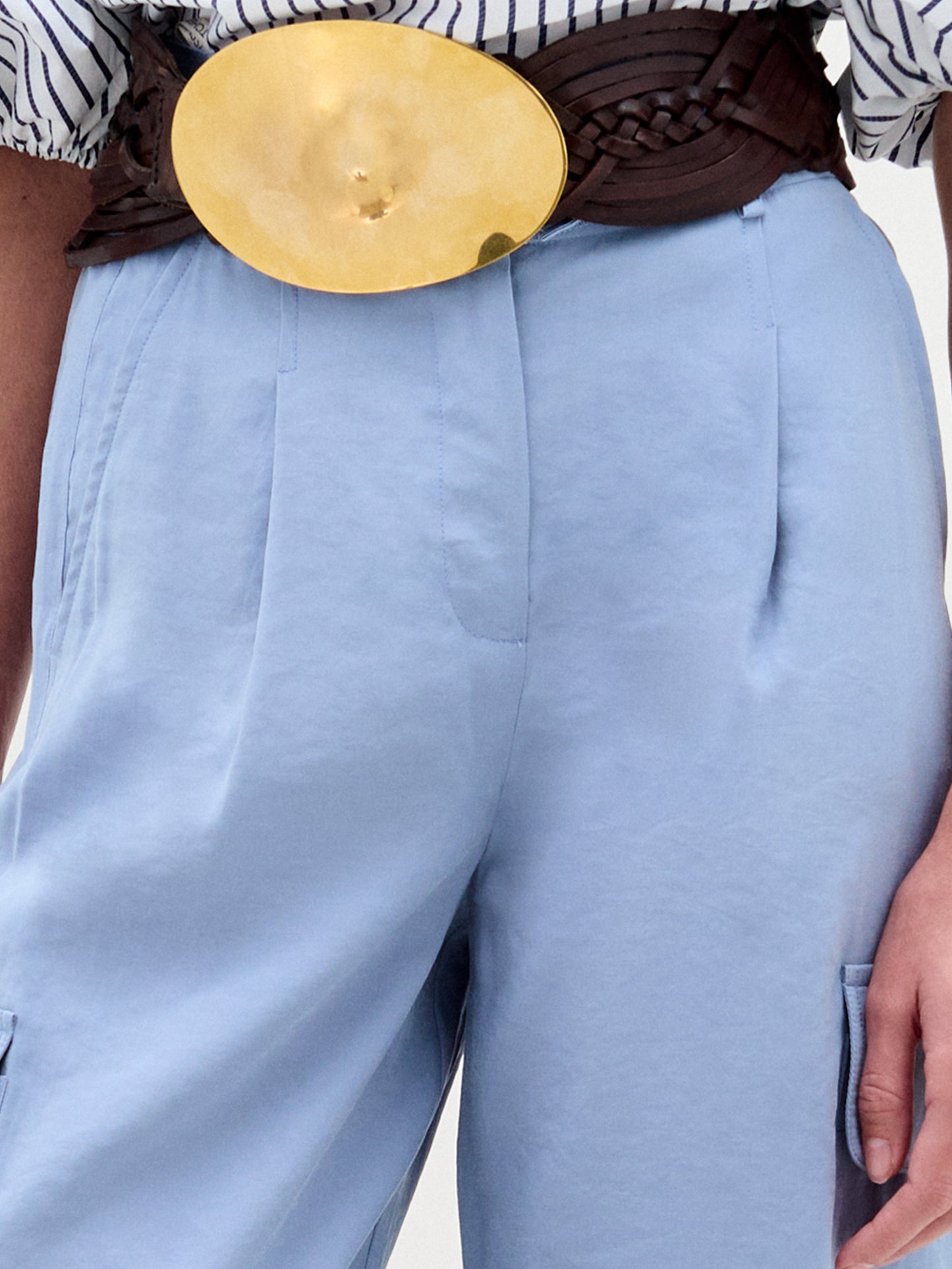 A person wearing SVT DESIGNS' Darnell Pant in light blue, paired with a wide dark brown woven belt featuring an oversized gold oval buckle. The black and white striped top is partially visible, with the hand resting at the person's side.