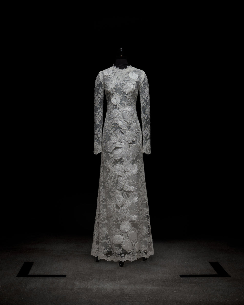 The Dava Dress by SVT DESIGNS is a full-length, long-sleeved gown with intricate lace patterns and floral designs from the high neckline to the hem, showcased on a black mannequin against a dark background.